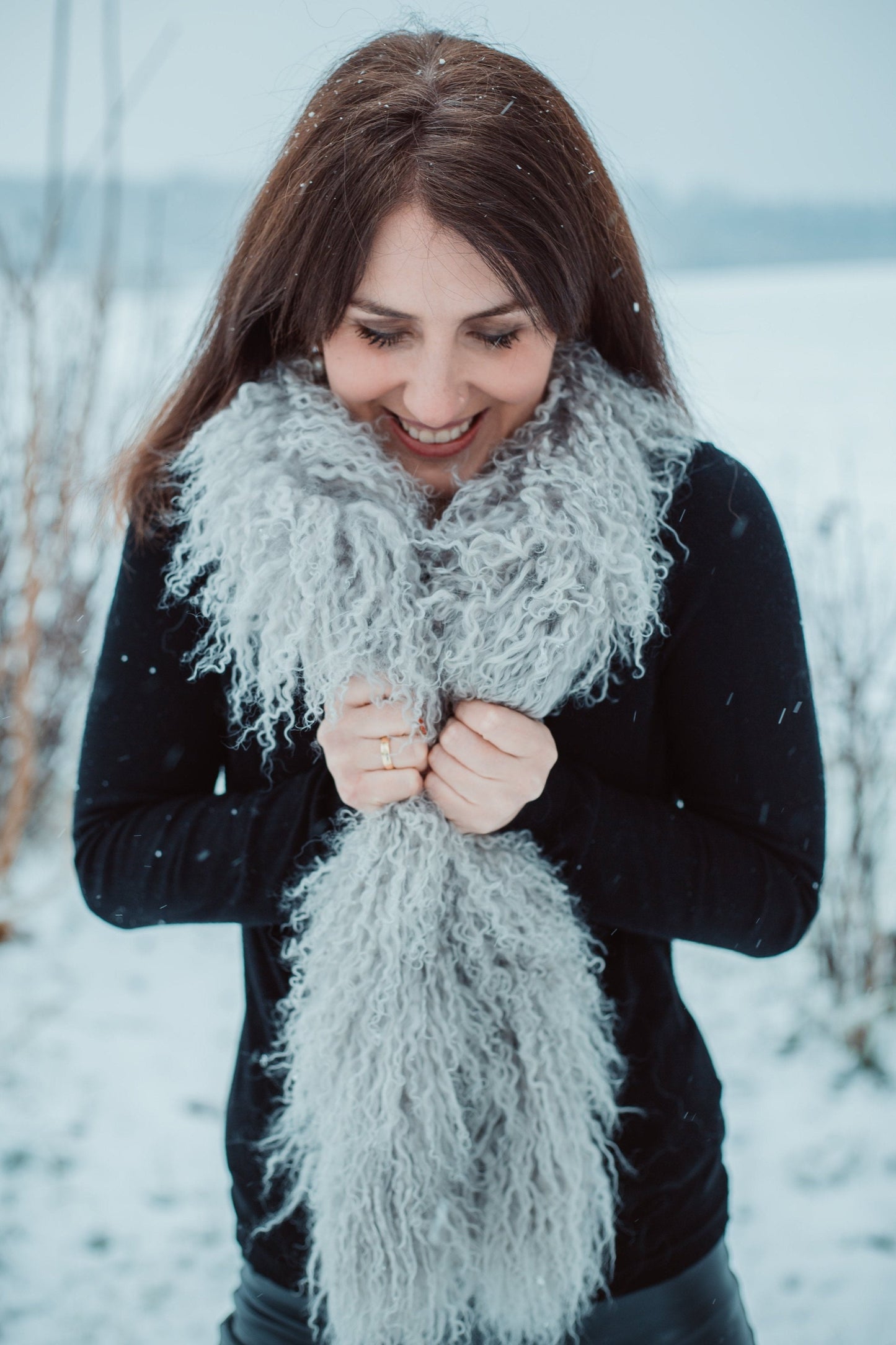Eine Person mit langen dunklen Haaren steht in einer verschneiten Landschaft. Sie trägt ein schwarzes Oberteil, Lederhosen und einen flauschigen Tibet Lammfell Schal. Sie hält den Schal in der Mitte und blickt nach unten. In der Luft sind Schneeflocken zu sehen. Im Hintergrund sind Bäume und ein riesiges Schneefeld zu sehen.