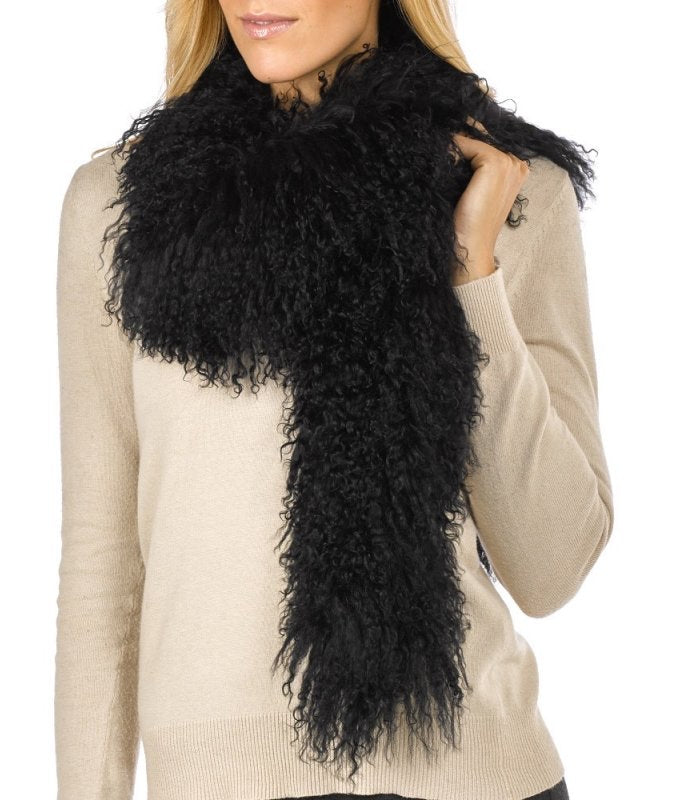 Eine Frau in einem schwarzen Langarmshirt und einer dunklen Hose präsentiert einen langen, schwarzen, Tibetlamm Schal Boa / Lammfell Schal. Der Schal wirkt weich und dick. Der Schal umwickelt mit einer Seite ihren Hals und fällt auf der anderen Seiten bis über ihre Taille. Der Hintergrund ist schlicht weiß.