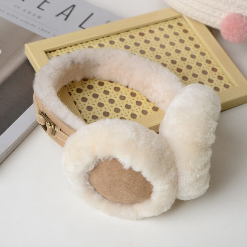 Ein Paar creme, flauschige Ohrenwärmer aus Lammfell. Die Ohrenwärmer sind mit zwei goldenen Accessories versehen und werden vor einem schlichten weißen Hintergrund auf einem Holzbrett und einem Magazin präsentiert.