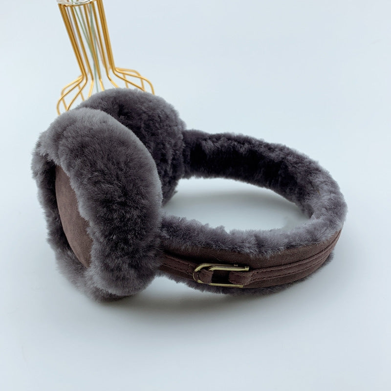 Ein Paar dunkelgrau, flauschige Ohrenwärmer aus Lammfell. Die Ohrenwärmer sind mit zwei goldenen Accessories versehen und werden vor einem schlichten weißen Hintergrund mit einer goldenen Metallvase präsentiert.