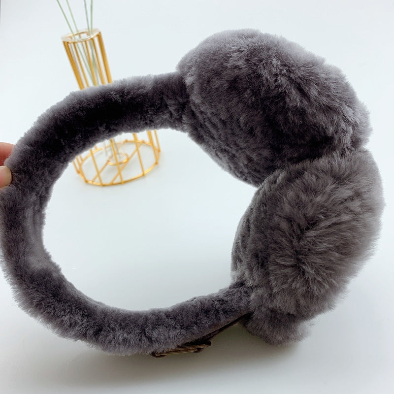 Ein Paar dunkelgrau, flauschige Ohrenwärmer aus Lammfell von vorne. Die Ohrenwärmer sind mit zwei goldenen Accessories versehen und werden vor einem schlichten weißen Hintergrund mit einer goldenen Metallvase präsentiert.