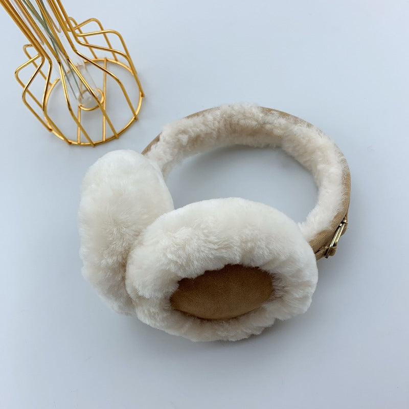 Ein Paar creme, flauschige Ohrenwärmer aus Lammfell. Die Ohrenwärmer sind mit zwei goldenen Accessories versehen und werden vor einem schlichten weißen Hintergrund mit einer goldenen Metallvase präsentiert.