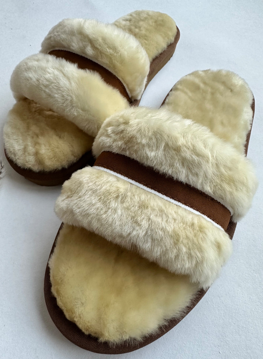 Ein Paar kuschelige „Schlappen in Beige/Creme“ von LIRA Deko, mit cremefarbenen Kunstfellriemen und gepolsterten braunen Sohlen. Diese Hausschuhe haben vorne zwei breite Riemen, die für Komfort und Wärme sorgen. Sie sind auf einem neutralen, hellen Untergrund platziert.