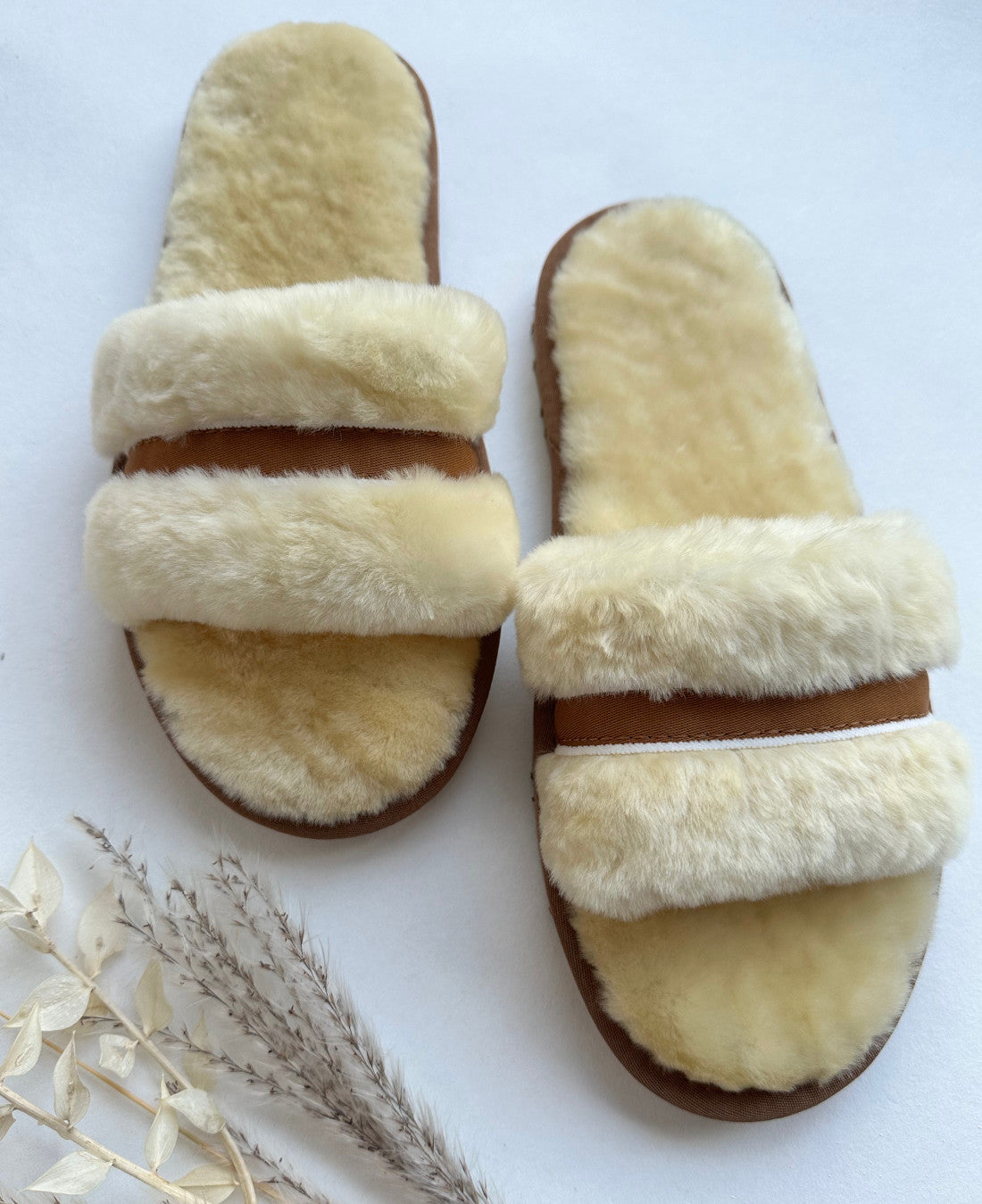 Ein Paar Schlappen von LIRA Deko in Beige/Creme mit kuscheligem, plüschigem Fellfutter aus echtem Lammfell und strapazierfähigen braunen Sohlen wird vor einem weißen Hintergrund präsentiert. Zu den Hausschuhen gehören zwei getrocknete Pflanzen: eine mit kleinen hellen Blättern und die andere mit zarten, federartigen Wedeln.