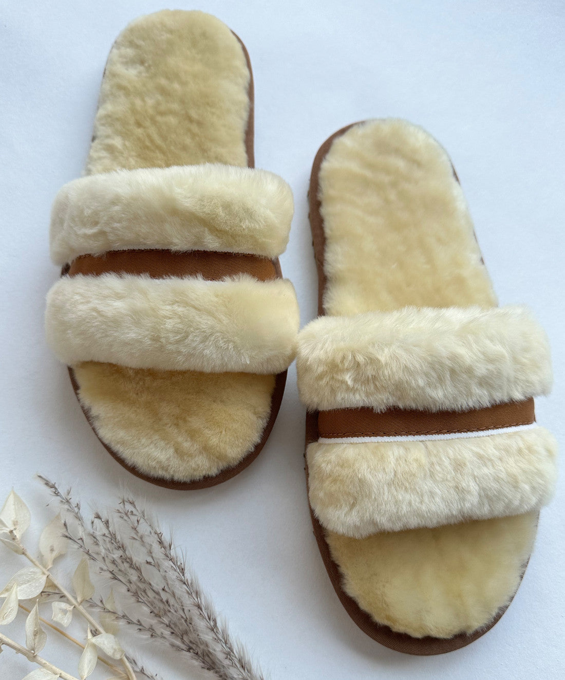 Ein Paar gemütliche, flauschige LIRA Deko Schlappen in Beige/Creme mit hellbraunen Sohlen und hellbeigen Kunstfellriemen. Die kuschlig und einladend wirkenden Pantoffeln stehen auf einem neutralen Hintergrund mit einigen getrockneten Pflanzenstängeln in der Ecke und sorgen für einen weichen und behaglichen Look.