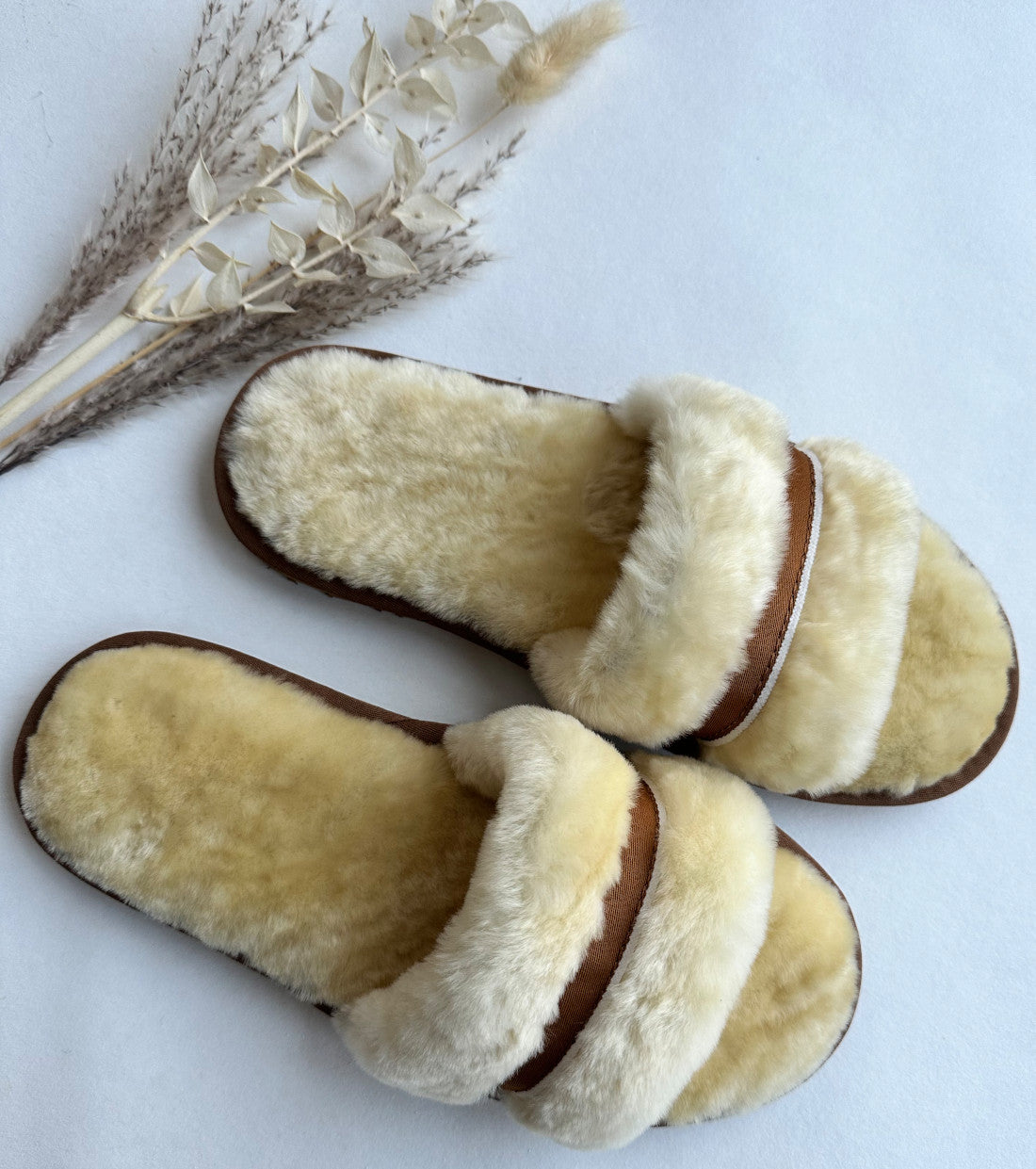 Ein Paar Schlappen in Beige/Creme von LIRA Deko aus echtem Lammfell mit offener Spitze und einem einzelnen Riemen über der Oberseite stehen auf einer weißen Oberfläche. Neben diesen Kuschlig-Sandalen steht ein kleines Arrangement aus getrockneten Gräsern und Weizenhalmen.