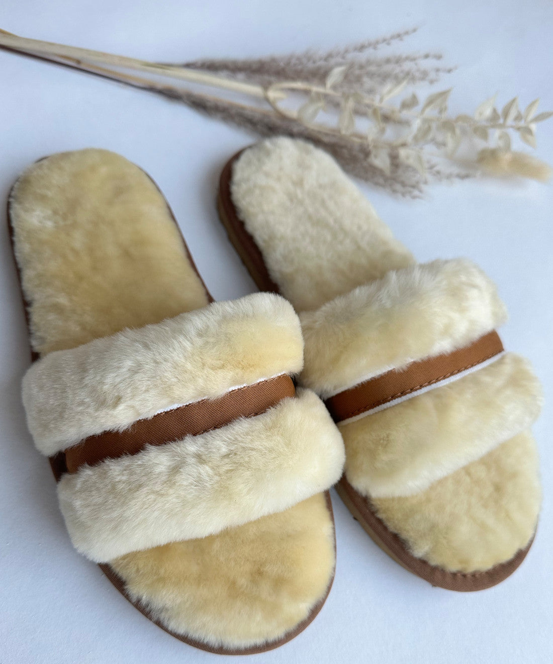Ein Paar Schlappen in Beige/Creme von LIRA Deko, mit kuschliger Fellfütterung aus echtem Lammfell und ergänzt durch braune Lederriemen, liegt auf einer weißen Unterlage. Getrocknete Weizenhalme sind kunstvoll neben den Schlappen arrangiert und schaffen eine warme und heimelige Atmosphäre.