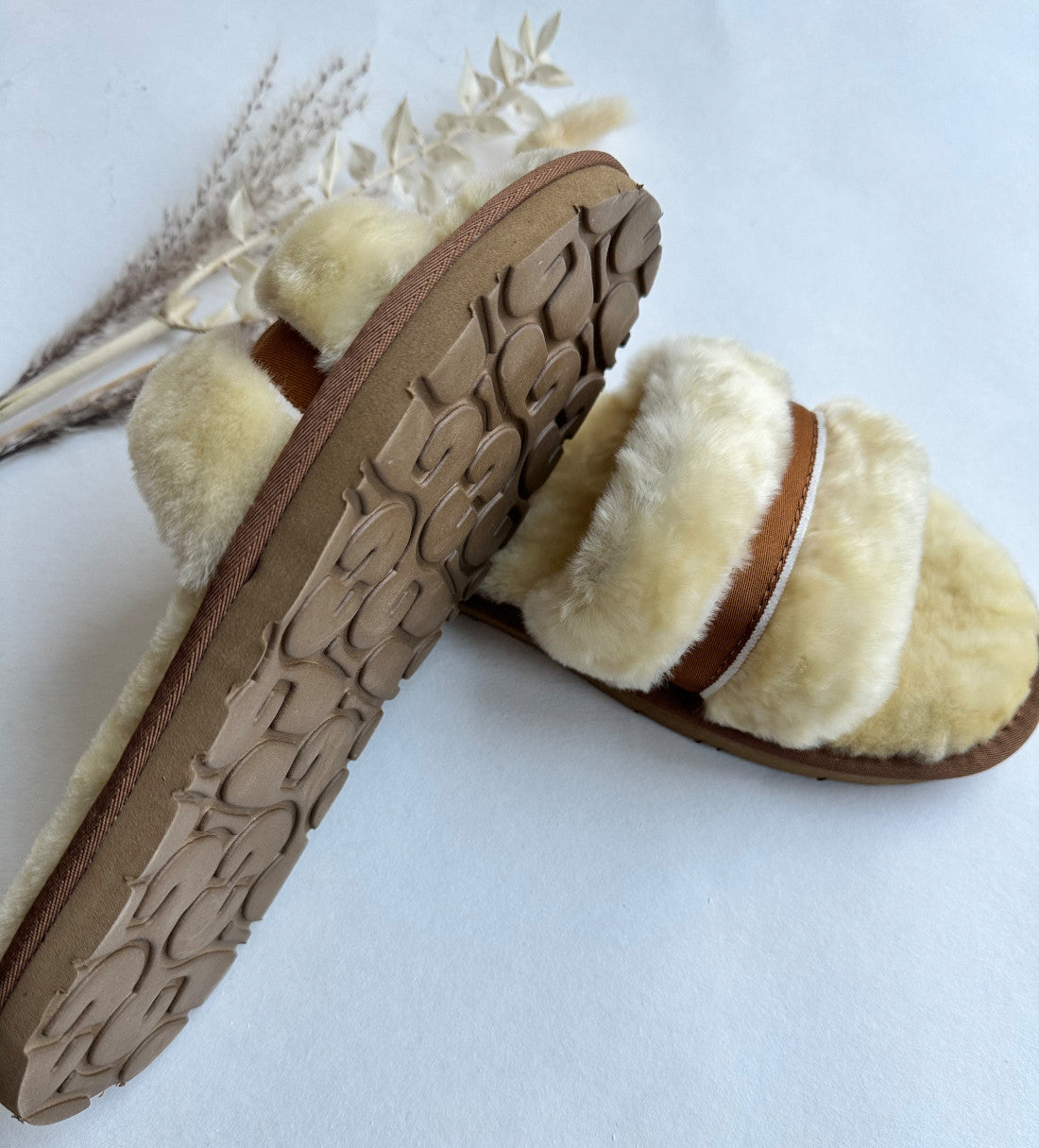 Ein Paar Schlappen in Beige/Creme von LIRA Deko wird auf einer weißen Fläche präsentiert. Diese kuscheligen, pelzgefütterten Hausschuhe aus echtem Lammfell haben flauschige cremefarbene Riemen und hellbraune Sohlen. Sie sind schräg angeordnet, einer überlappt den anderen leicht. Im Hintergrund verleihen getrocknete Gräser und Blumen einen Hauch natürlicher Eleganz.